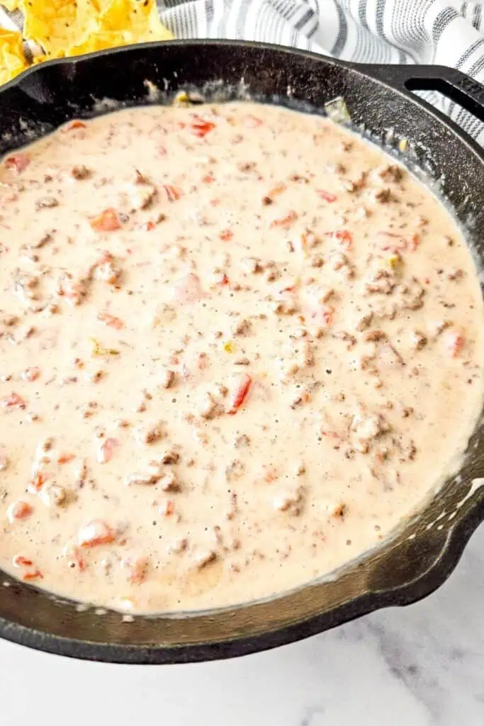 A cast iron skillet filled with creamy Queso Blanco Rotel Dip containing ground meat and diced tomatoes.