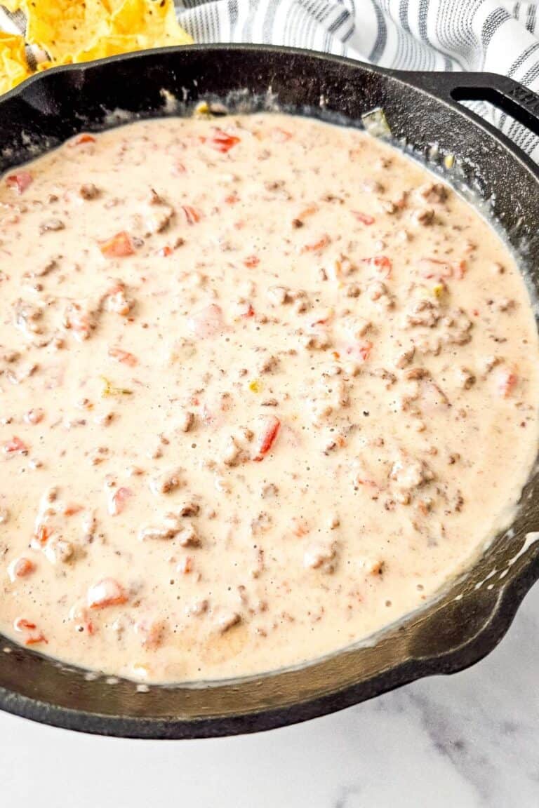 A cast iron skillet filled with creamy Queso Blanco Rotel Dip containing ground meat and diced tomatoes.
