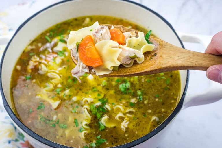A wooden spoon holds chicken noodle soup with carrots and herbs above a pot of soup.