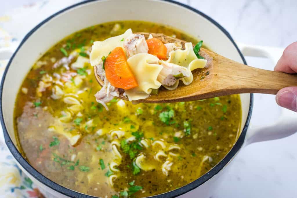 Wooden spoon with Rotisserie chicken noodle soup, featuring noodles, chicken pieces, and carrots, over a pot filled with broth and garnished with herbs.
