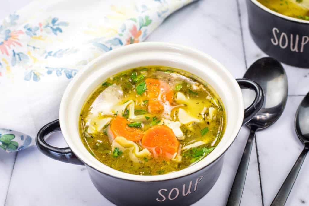 A black bowl of rotisserie chicken noodle soup, containing shredded chicken, egg noodles, sliced carrots, and fresh herbs in a golden broth.