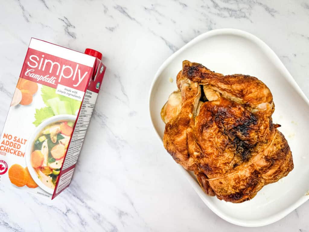 A roasted chicken on a white dish sits beside a carton of Campbell's Simply chicken broth on a marble surface.