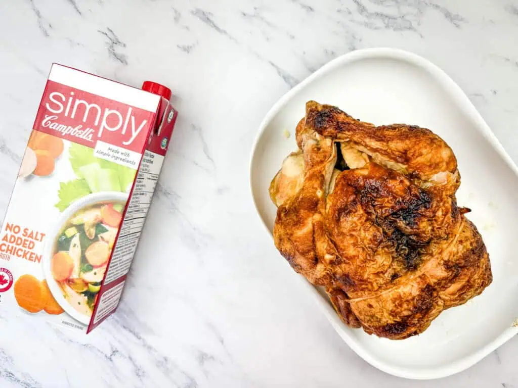 A roasted chicken on a white dish sits beside a carton of Campbell's Simply chicken broth on a marble surface.