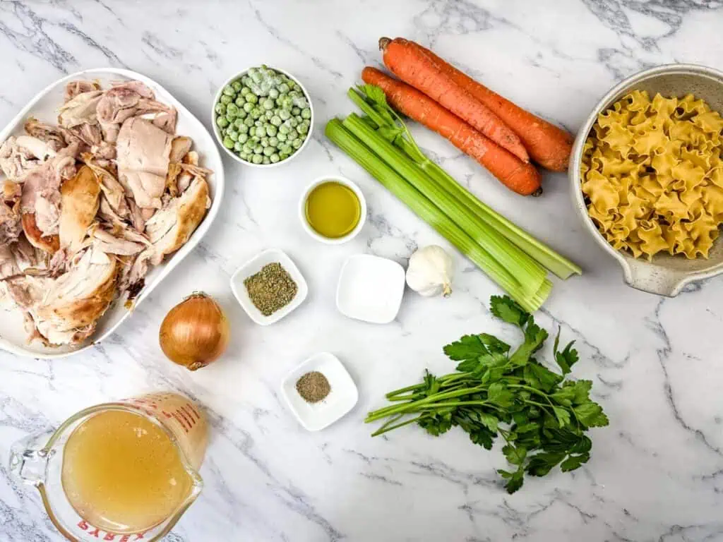 Ingredients on a marble surface include shredded chicken, frozen peas, carrots, celery, lasagna noodles, broth, onion, olive oil, garlic, parsley, and small bowls of spices.