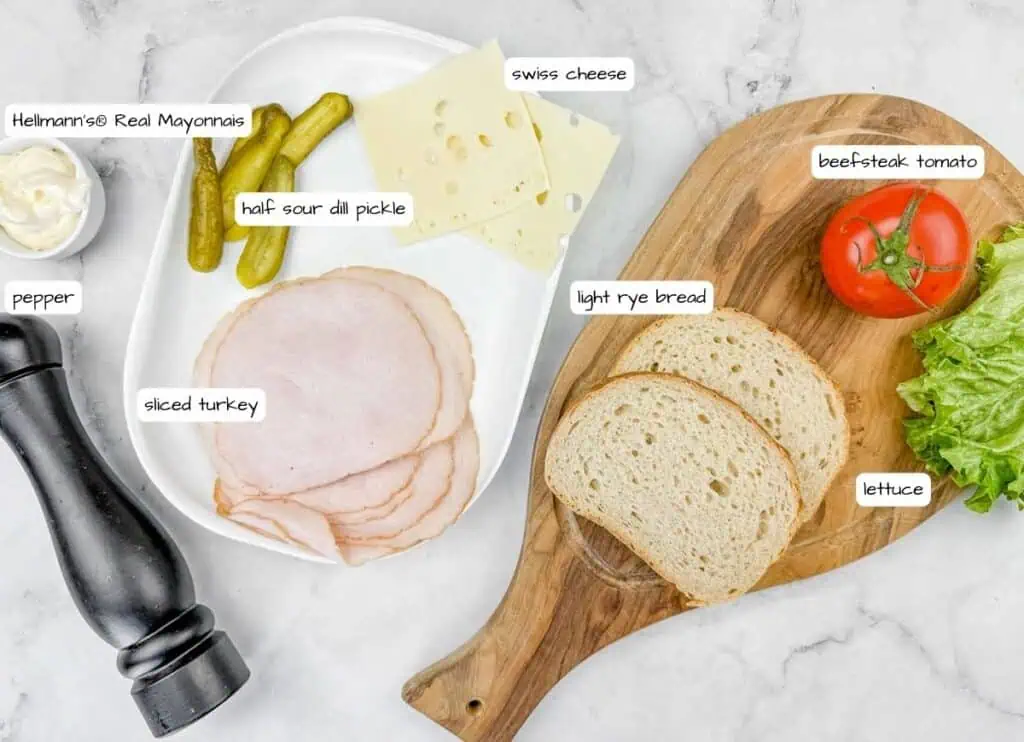 Labeled ingredients for What She's Having a Sandwich on a wooden board and plate on top of white marble countertop.