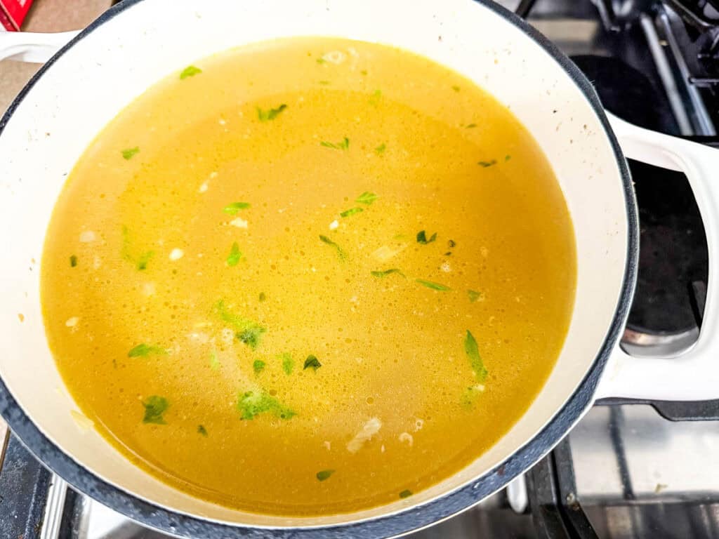 A white pot containing yellow broth with visible herbs and pieces on a stove.