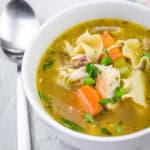 A white bowl filled with rotisserie chicken noodle soup, featuring shredded chicken, egg noodles, carrots, celery, peas, and fresh herbs in a golden broth. A spoon rests beside the bowl on a textured surface.