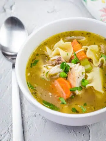 A white bowl filled with rotisserie chicken noodle soup, featuring shredded chicken, egg noodles, carrots, celery, peas, and fresh herbs in a golden broth. A spoon rests beside the bowl on a textured surface.