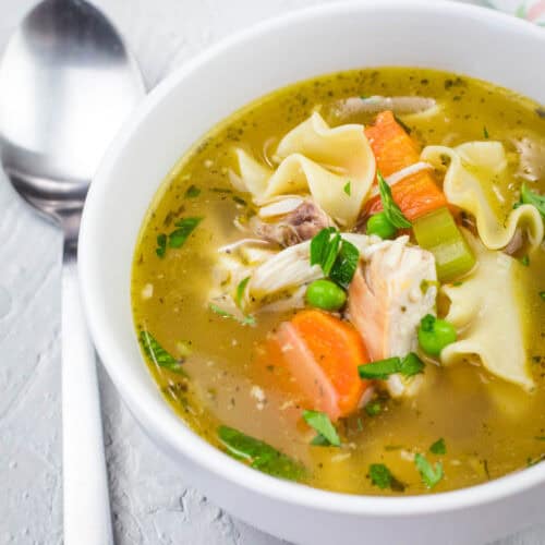 A white bowl filled with rotisserie chicken noodle soup, featuring shredded chicken, egg noodles, carrots, celery, peas, and fresh herbs in a golden broth. A spoon rests beside the bowl on a textured surface.