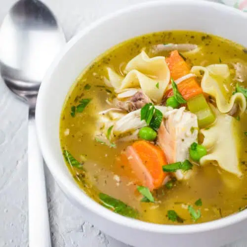 A white bowl filled with rotisserie chicken noodle soup, featuring shredded chicken, egg noodles, carrots, celery, peas, and fresh herbs in a golden broth. A spoon rests beside the bowl on a textured surface.