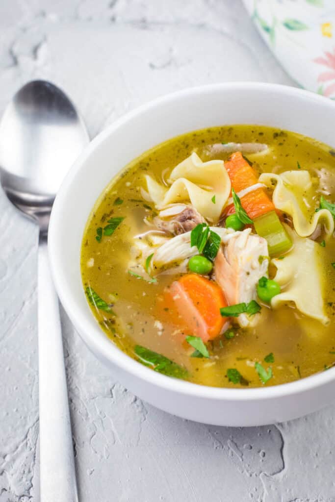 A white bowl filled with rotisserie chicken noodle soup, featuring shredded chicken, egg noodles, carrots, celery, peas, and fresh herbs in a golden broth. A spoon rests beside the bowl on a textured surface.