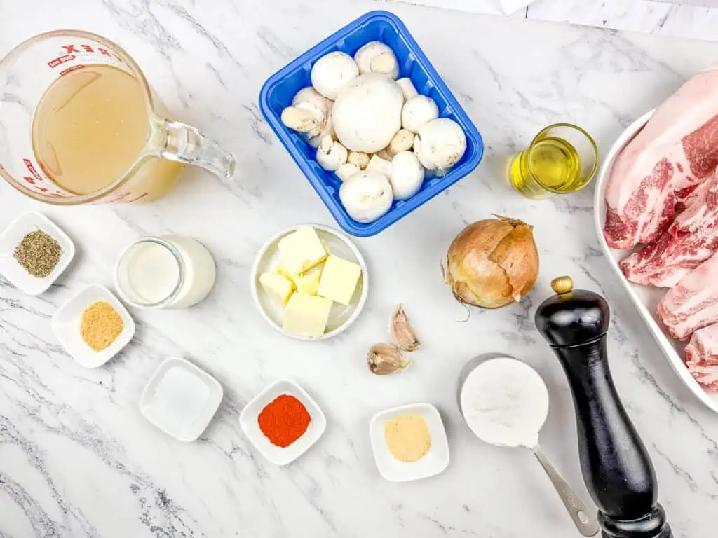 Ingredients for Smothered Pork Chops with Onions & Mushrooms on a marble countertop including mushrooms, pork, broth, onion, butter, cream, olive oil, spices, flour, garlic, pepper grinder, and measuring cup.