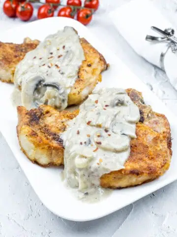 Two Smothered Pork Chops with Onions & Mushrooms on a white plate. Cherry tomatoes and a napkin with a silver holder are in the background.