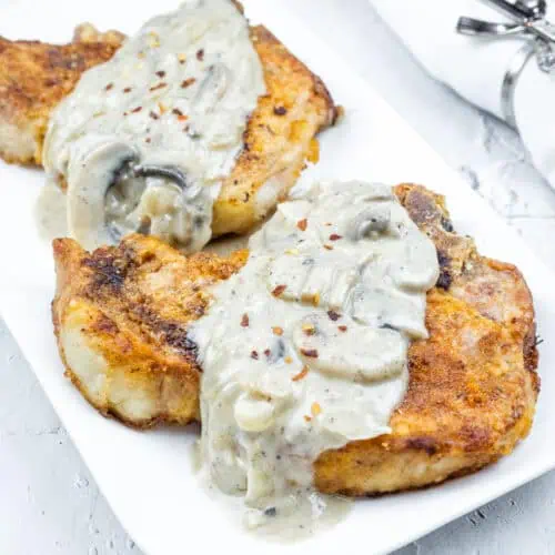 Two Smothered Pork Chops with Onions & Mushrooms on a white plate. Cherry tomatoes and a napkin with a silver holder are in the background.