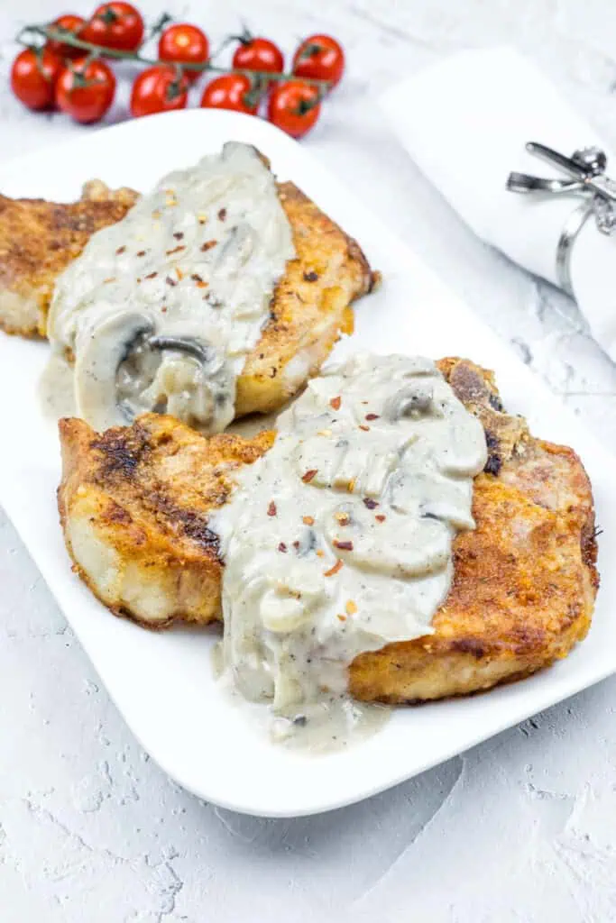 Two Smothered Pork Chops with Onions & Mushrooms on a white plate. Cherry tomatoes and a napkin with a silver holder are in the background.