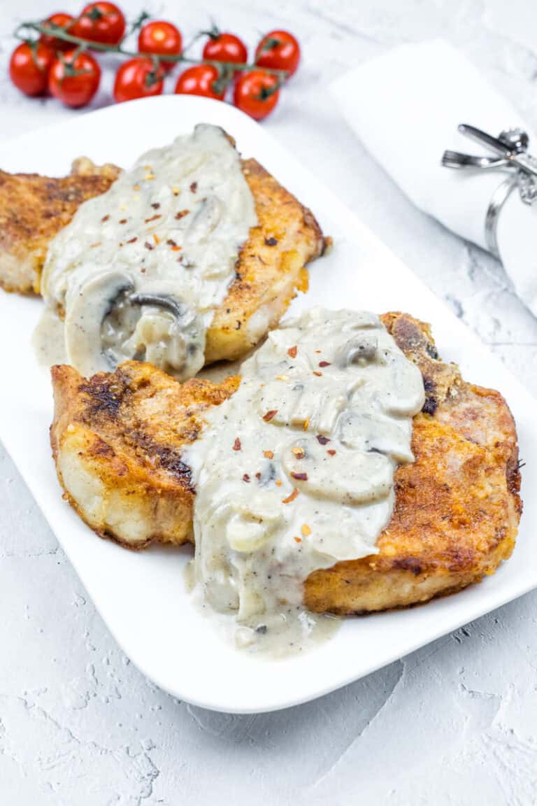 Two Smothered Pork Chops with Onions & Mushrooms on a white plate. Cherry tomatoes and a napkin with a silver holder are in the background.