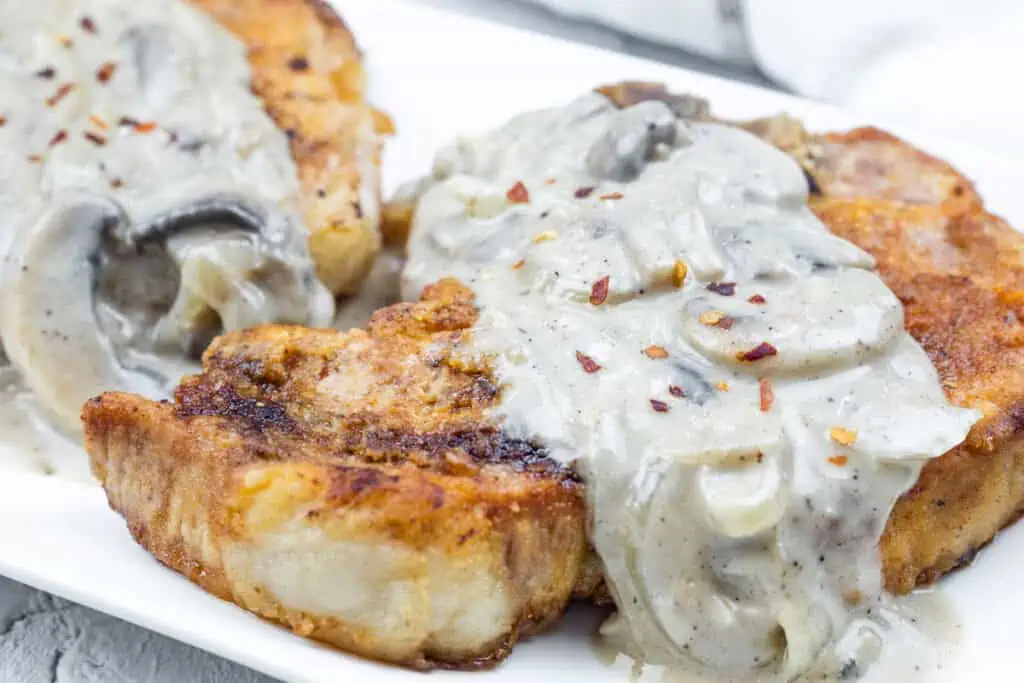 Two Smothered Pork Chops with Onions & Mushrooms, garnished with crushed red pepper, served on a white plate.
