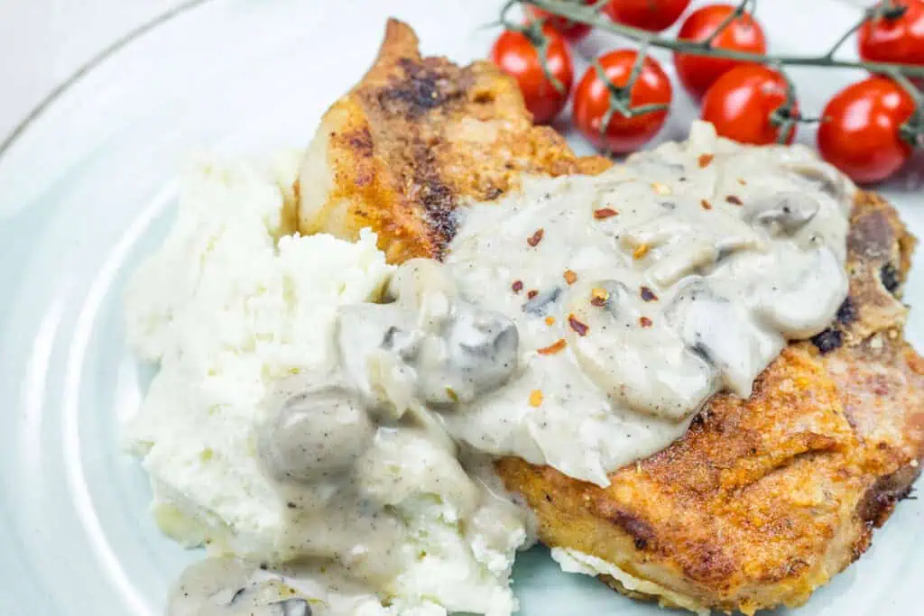 Plate of Smothered Pork Chops with Onions & Mushrooms, served with mashed potatoes and a side of cherry tomatoes.