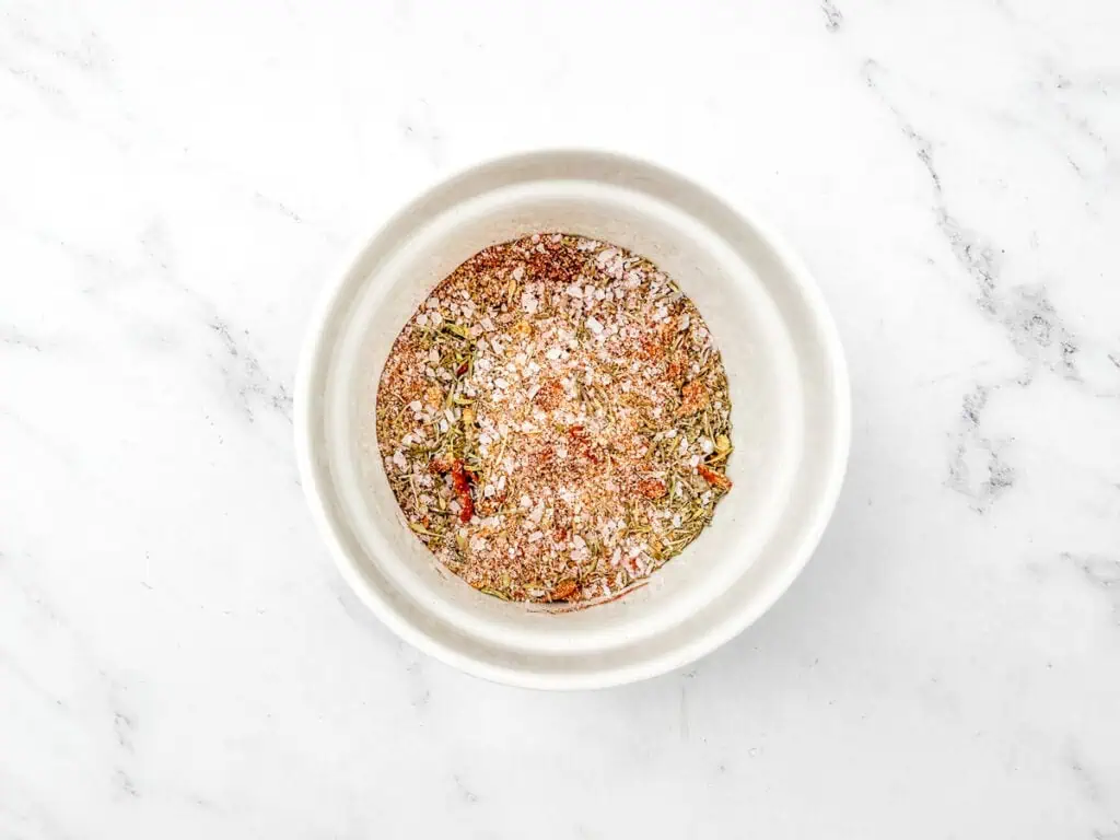 A white bowl containing a mix of various spices and herbs on a white marble surface.
