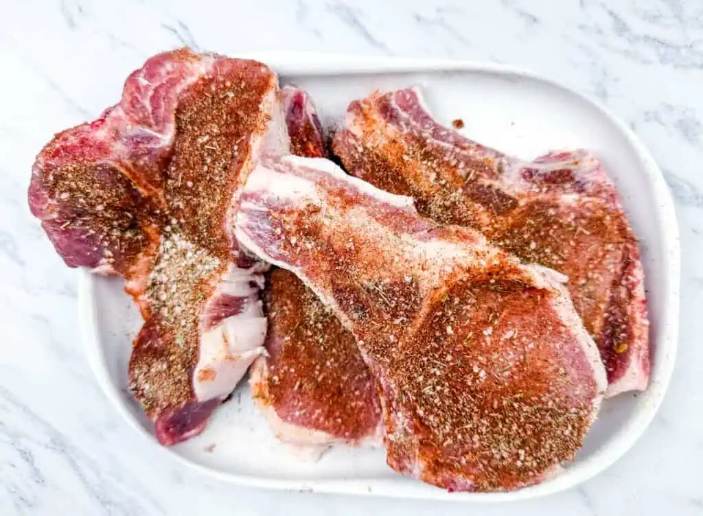 Three raw, seasoned pork chops on a white plate placed on a marble surface.