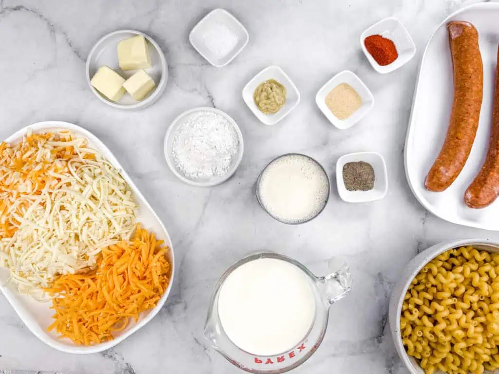 Ingredients on a countertop including shredded cheese, macaroni, milk, sausages, butter, and spices in small dishes.