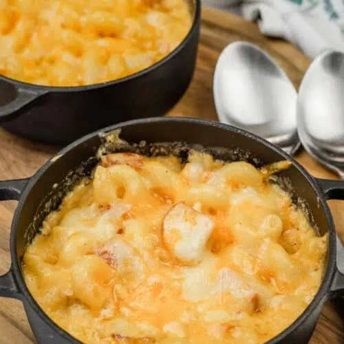 Two small cast iron pots filled with baked macaroni and cheese, placed on a wooden board, with two spoons beside them.