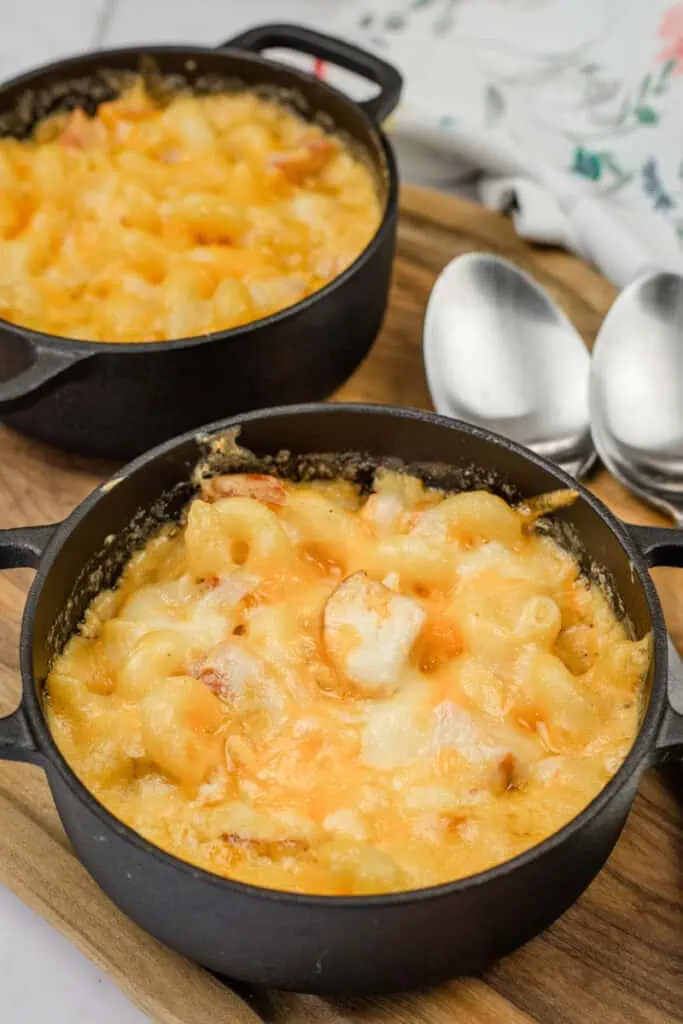 Two small cast iron pots filled with baked macaroni and cheese, placed on a wooden board, with two spoons beside them.