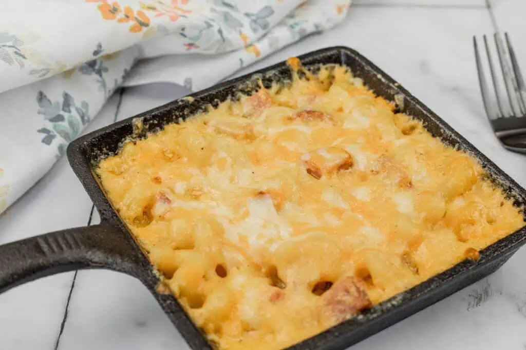 A small black cast iron pan filled with baked macaroni and cheese topped with melted cheese, placed on a marble surface next to a floral-patterned cloth and a fork.