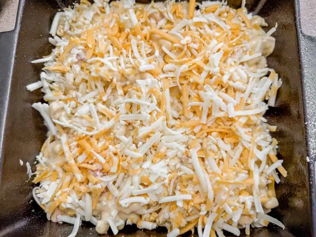 A square baking dish filled with shredded cheese mix on top of a layered dish, ready for baking.