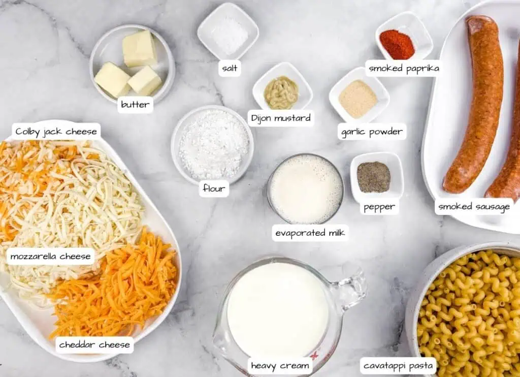 Ingredients labeled for Tini’s Mac & Cheese with a Twist on a marble surface: Colby jack, mozzarella, and cheddar cheese, butter, flour, evaporated milk, heavy cream, smoked paprika, Dijon mustard, garlic powder, pepper, smoked sausage, pasta.