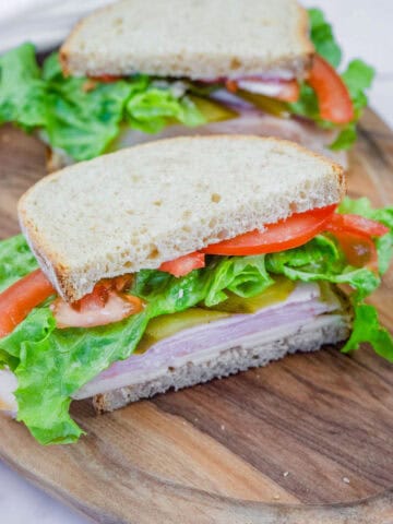 A freshly made turkey sandwich on whole-grain bread, layered with lettuce, tomato slices, pickles, and cheese, served on a wooden cutting board. The sandwich is cut in half, revealing the colorful and fresh ingredients inside.