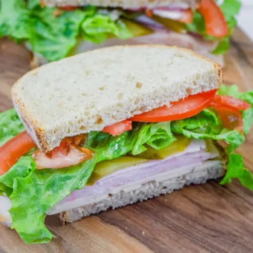A freshly made turkey sandwich on whole-grain bread, layered with lettuce, tomato slices, pickles, and cheese, served on a wooden cutting board. The sandwich is cut in half, revealing the colorful and fresh ingredients inside.