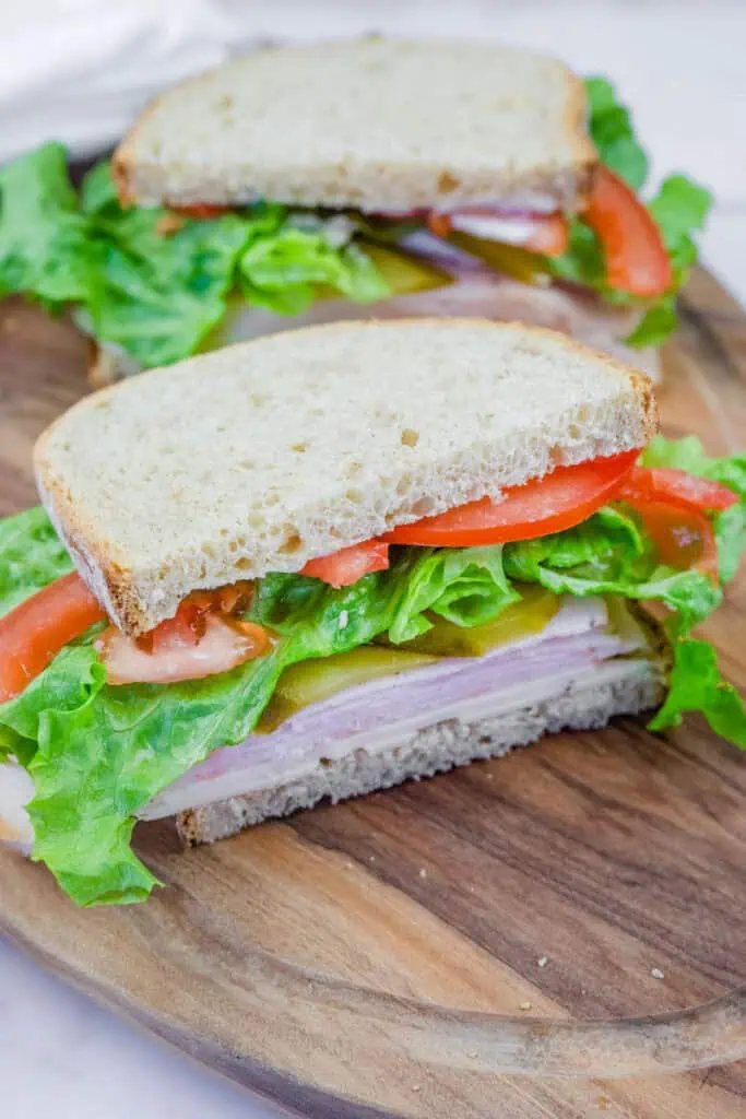 A freshly made turkey sandwich on whole-grain bread, layered with lettuce, tomato slices, pickles, and cheese, served on a wooden cutting board. The sandwich is cut in half, revealing the colorful and fresh ingredients inside.