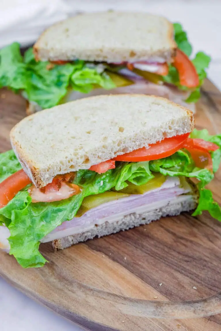 A freshly made turkey sandwich on whole-grain bread, layered with lettuce, tomato slices, pickles, and cheese, served on a wooden cutting board. The sandwich is cut in half, revealing the colorful and fresh ingredients inside.