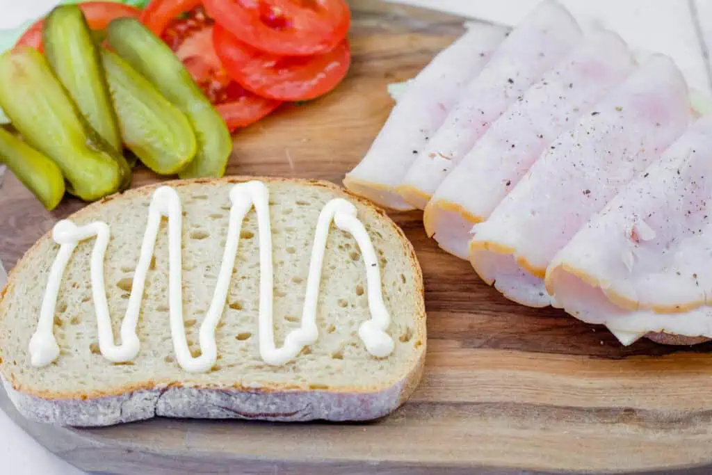 A close-up of sandwich ingredients arranged on a wooden cutting board. A slice of rustic bread with a zigzag drizzle of mayonnaise is in the foreground. To the right, several folded slices of turkey or ham are seasoned with black pepper. In the background, sliced pickles and fresh tomato slices are neatly arranged.