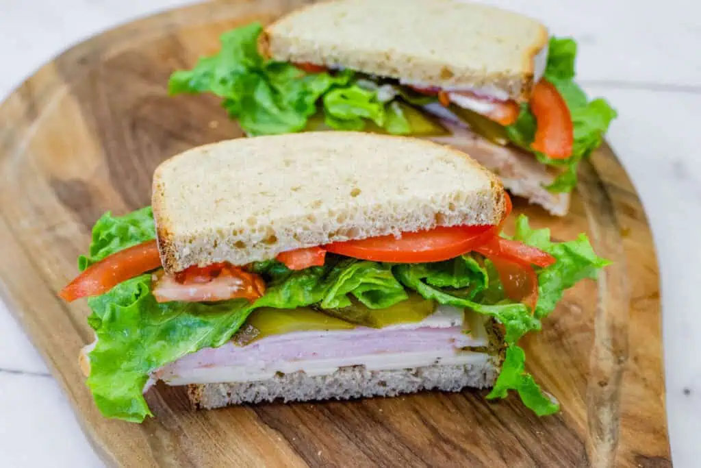 Turkey sandwich with lettuce, tomatoes, pickles, and cheese on whole-grain bread, cut in half and served on a wooden board.