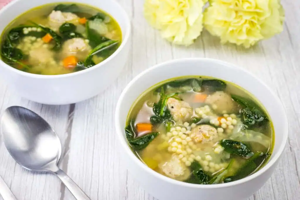 Two bowls of Italian Wedding Soup with spoon and flower on a white wooden table.