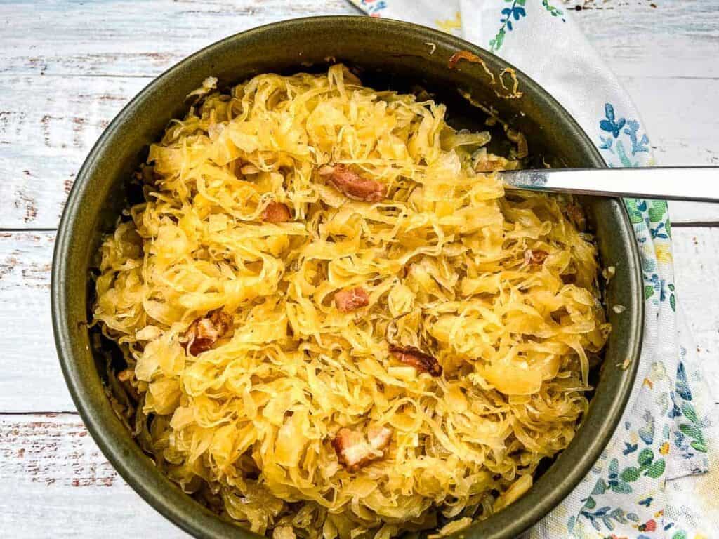 Bowl of cooked sauerkraut mixed with bacon pieces, with a spoon, on a white wooden surface beside a patterned napkin.
