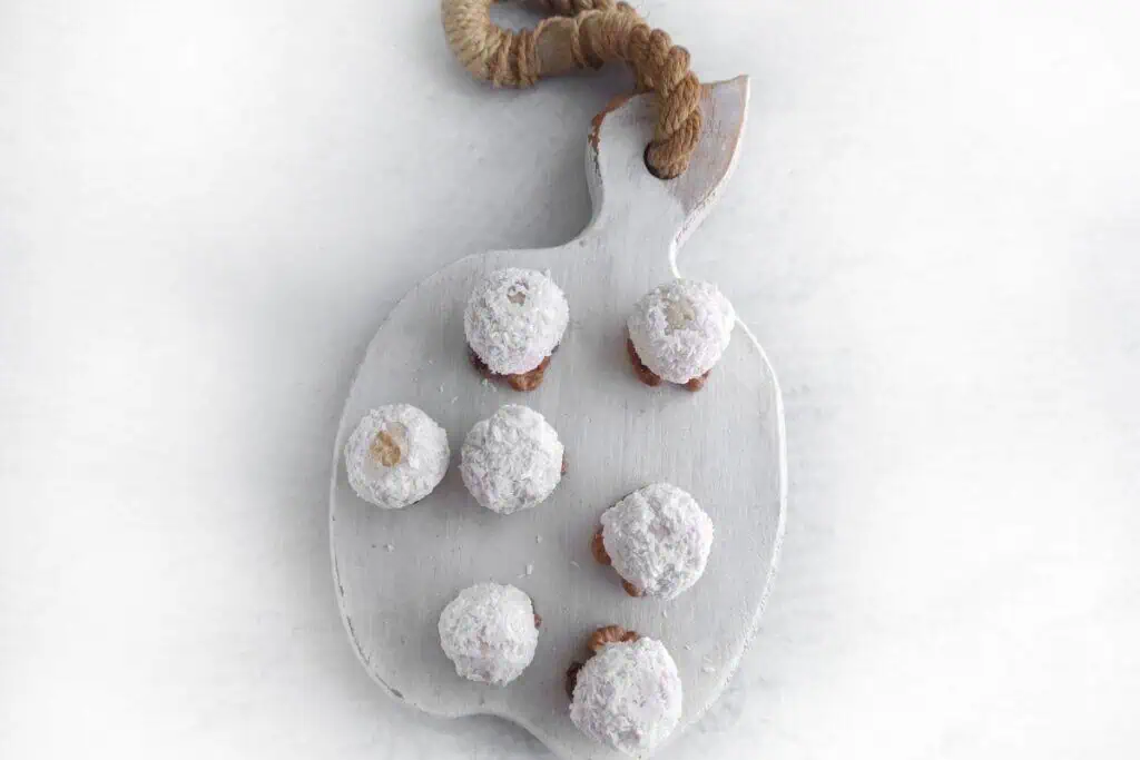 Six powdered sugar-covered cookies are arranged on a white cutting board with a rope handle, placed on a light surface.
