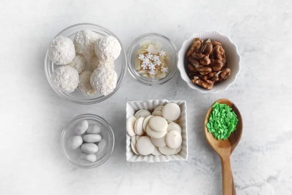 Assorted baking ingredients displayed on a table include coconut balls, almond slices, walnuts, white almonds, white chocolate buttons, and green sprinkles in a wooden spoon.