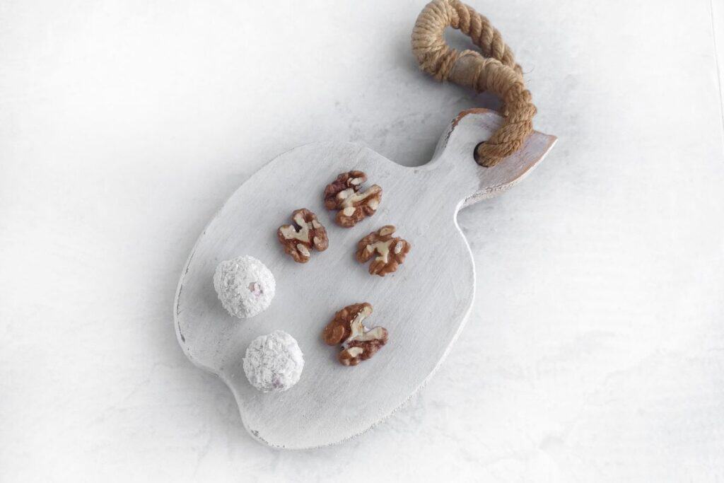 Sliced walnuts and two white powdered truffles on a rustic white cutting board with a rope handle, set against a light background.
