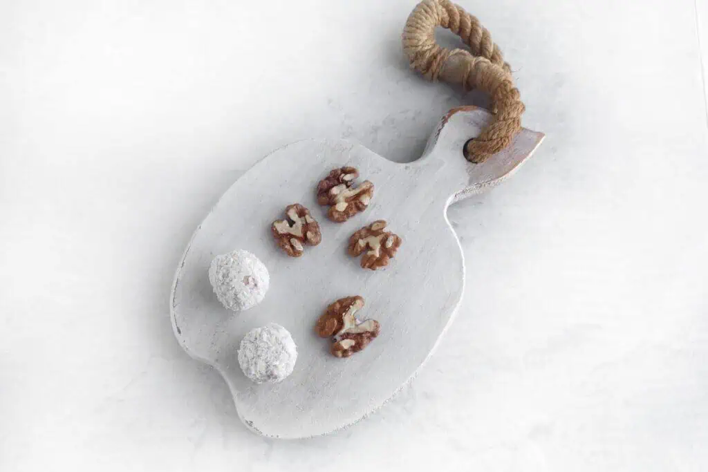 Sliced walnuts and two white powdered truffles on a rustic white cutting board with a rope handle, set against a light background.