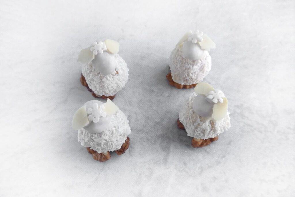 Four small, round confections resembling sheep, with white coconut fur, almond ears, and floral decorations on a light background.