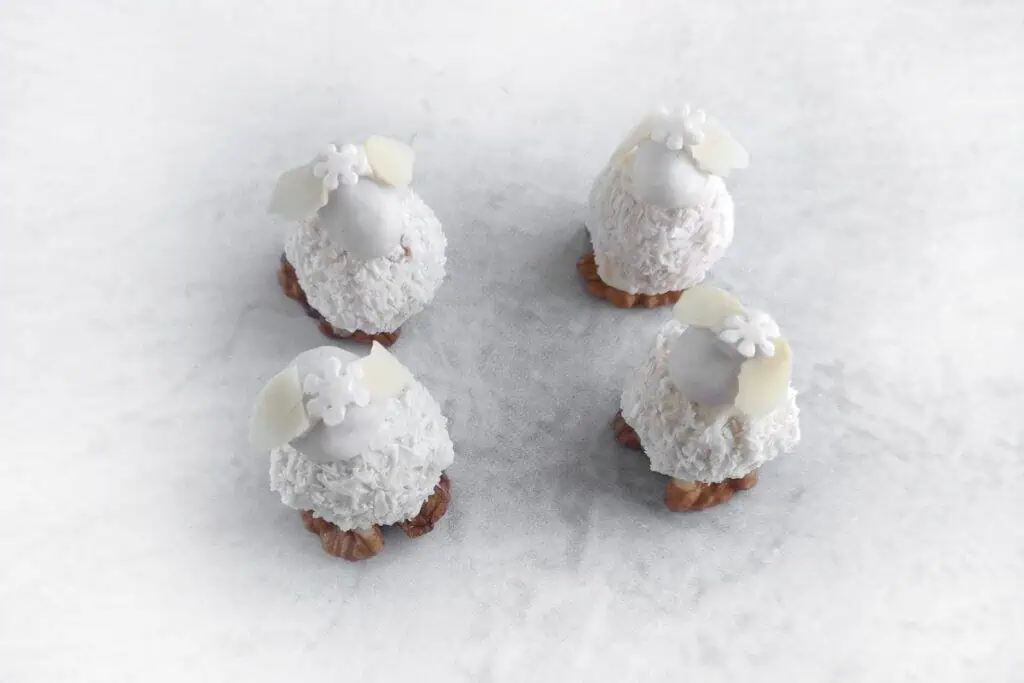 Four small, round confections resembling sheep, with white coconut fur, almond ears, and floral decorations on a light background.