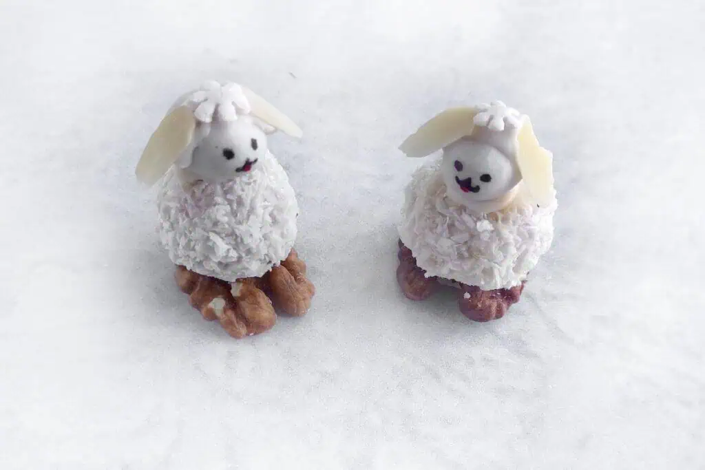 Two small sheep-shaped confections with white frosting, coconut flakes, and walnut legs on a white textured surface.
