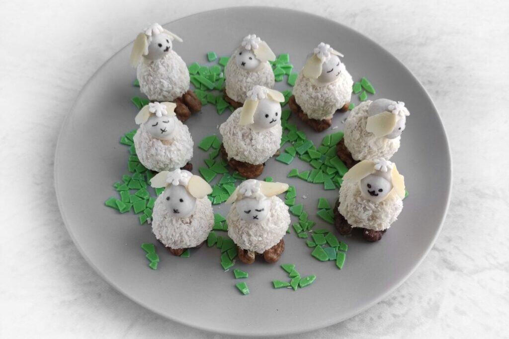 Plate with nine coconut-coated sheep-shaped desserts, each with almond ears and chocolate faces, surrounded by green chocolate shard decorations.
