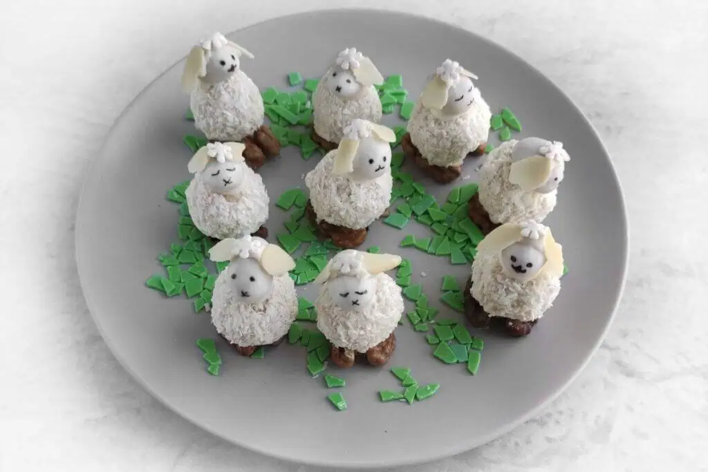 Plate with nine coconut-coated sheep-shaped desserts, each with almond ears and chocolate faces, surrounded by green chocolate shard decorations.