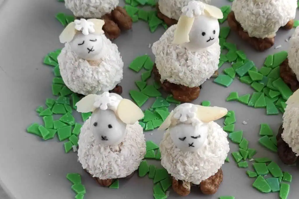 Cute sheep-shaped treats made with coconut flakes and icing on a plate, surrounded by green candy pieces.