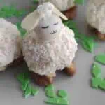 A dessert shaped like a sheep, featuring a coconut-covered body, almond head, and small eyes and mouth. It stands on walnuts with green candy pieces scattered around.