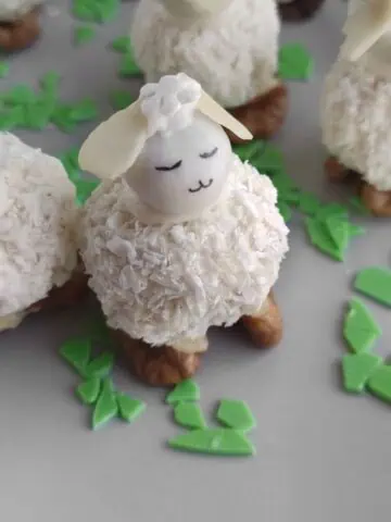 A dessert shaped like a sheep, featuring a coconut-covered body, almond head, and small eyes and mouth. It stands on walnuts with green candy pieces scattered around.